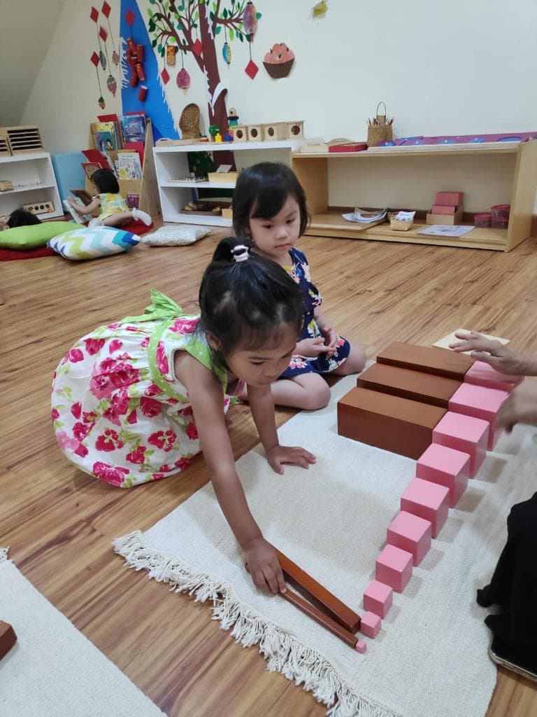 Dua anak menyusun balok pink tower dan brown stair Montessori di atas alas lantai