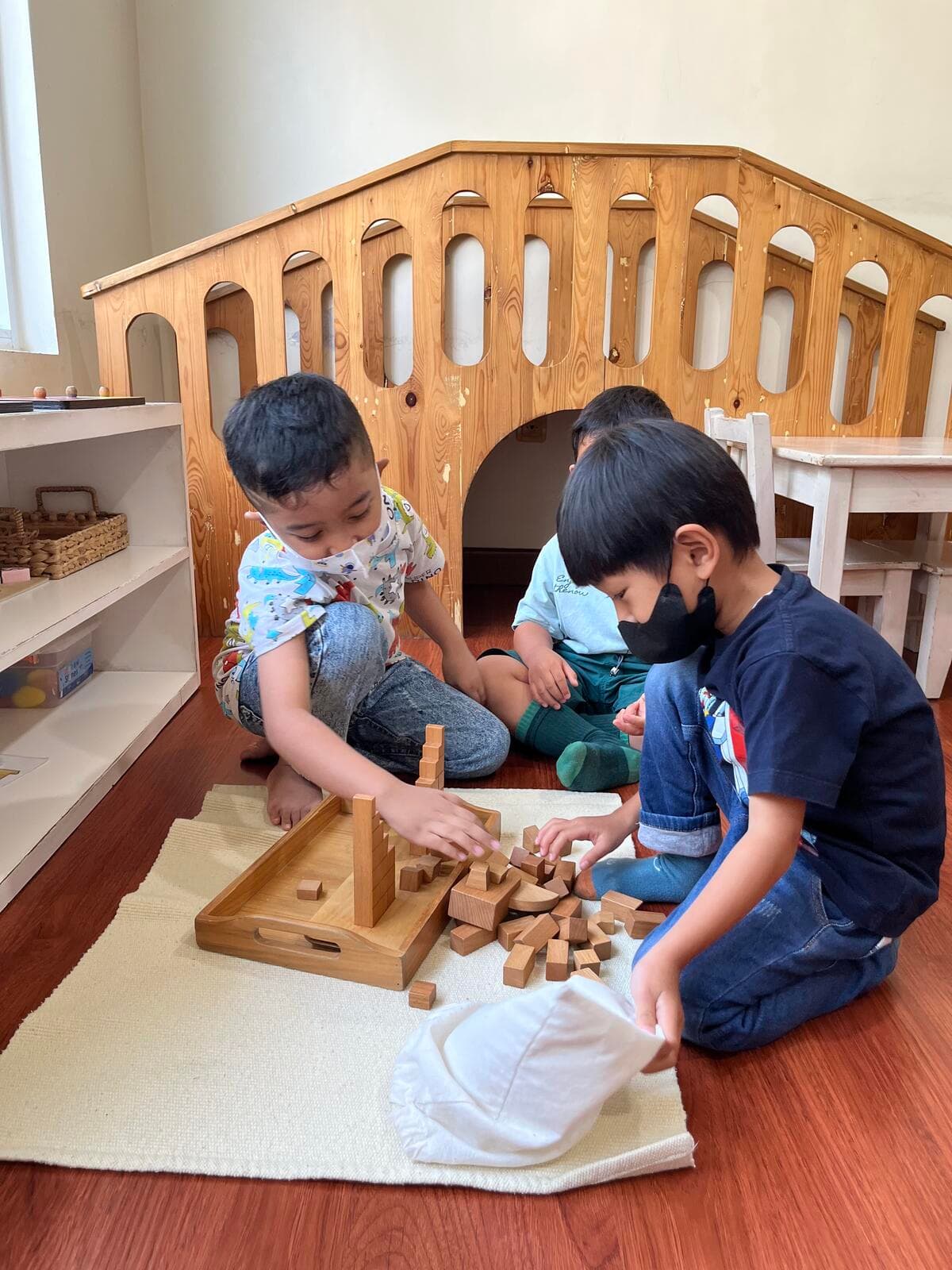 Two older boys sorting wooden blocks from a tray on a floor mat while a younger toddler watches, in front of a wooden climbing arch