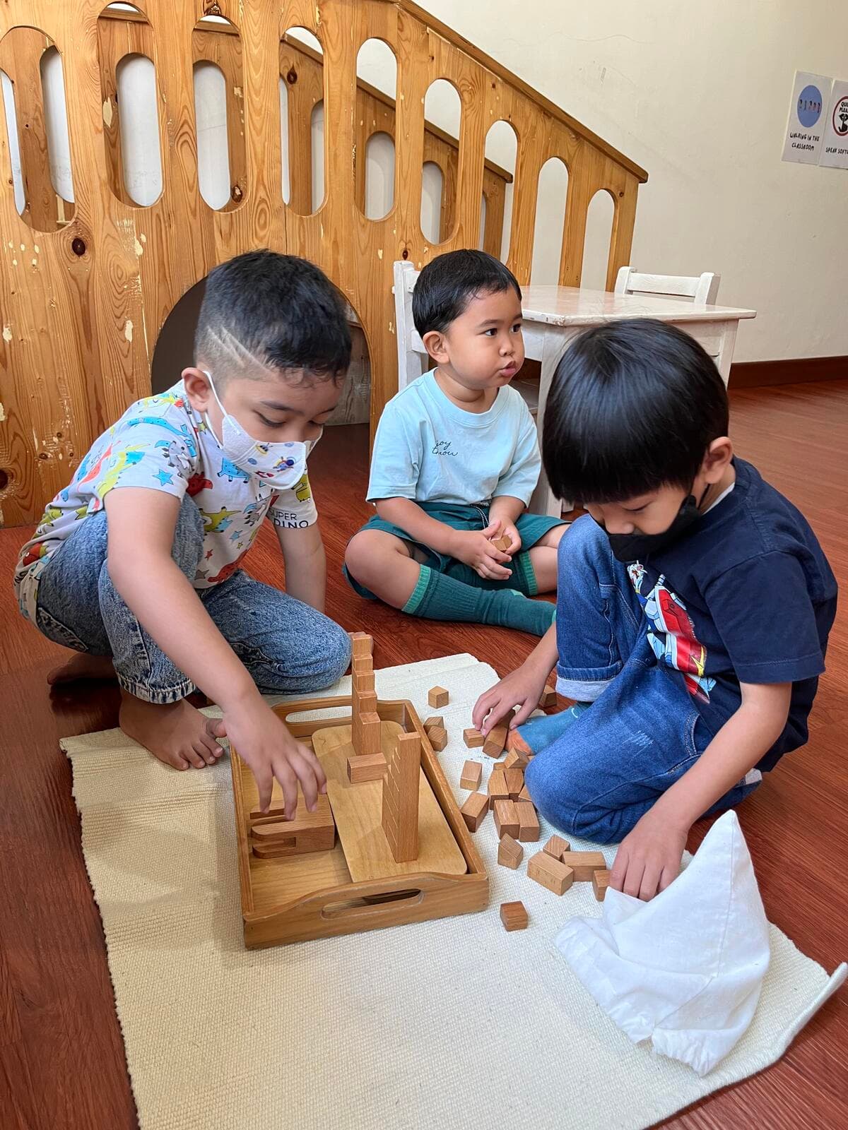 Two boys building a block tower in a wooden tray while a younger child observes, in front of a wooden climbing arch
