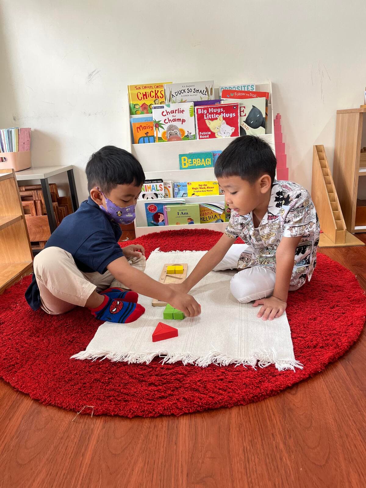Two boys arranging colorful geometric solids on a white work mat on a round red rug, with a bookshelf of picture books behind them