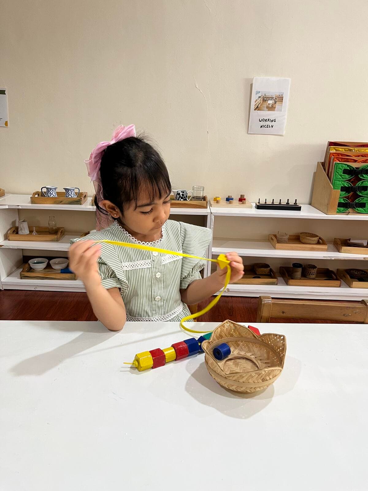 A girl with a pink headband threading colorful beads onto a yellow lace at a table, with practical life materials on shelves behind her
